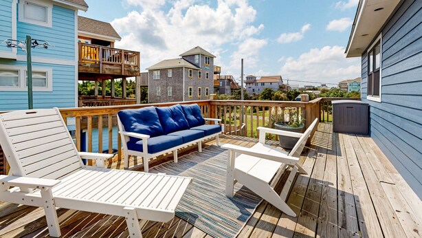 Una terraza de madera con sillas blancas y un sofá azul, con vistas a una zona residencial con casas y un cielo despejado.