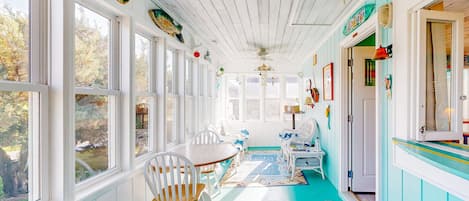 Une pièce baignée de lumière, avec un plafond en bois, des murs blancs et un sol de couleur turquoise. On y trouve une table à manger avec des chaises, une petite table blanche et un banc.
