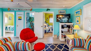 A brightly colored beach-themed living room with striped furniture, a red chair, and a sign reading "Welcome to our beach house."