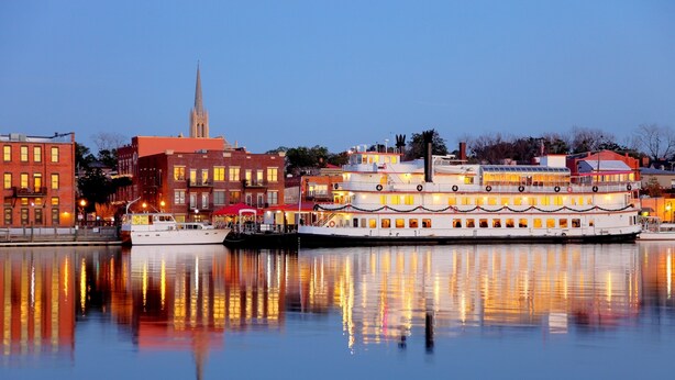 A multi-story riverboat with a white exterior and multiple decks, docked near a cityscape with buildings and a church spire. - Historical Riverfront Condo in Historic District, Highly Walkable Locale (Wilmington)