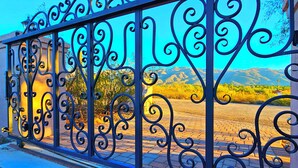 Una cerca de hierro forjado con diseños intrincados, que da paso a una zona pavimentada con vistas a un paisaje montañoso.