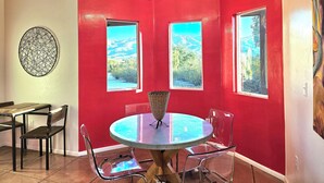 Un comedor con una mesa redonda, sillas transparentes y una araña. Hay tres ventanas con vistas a un paisaje montañoso.