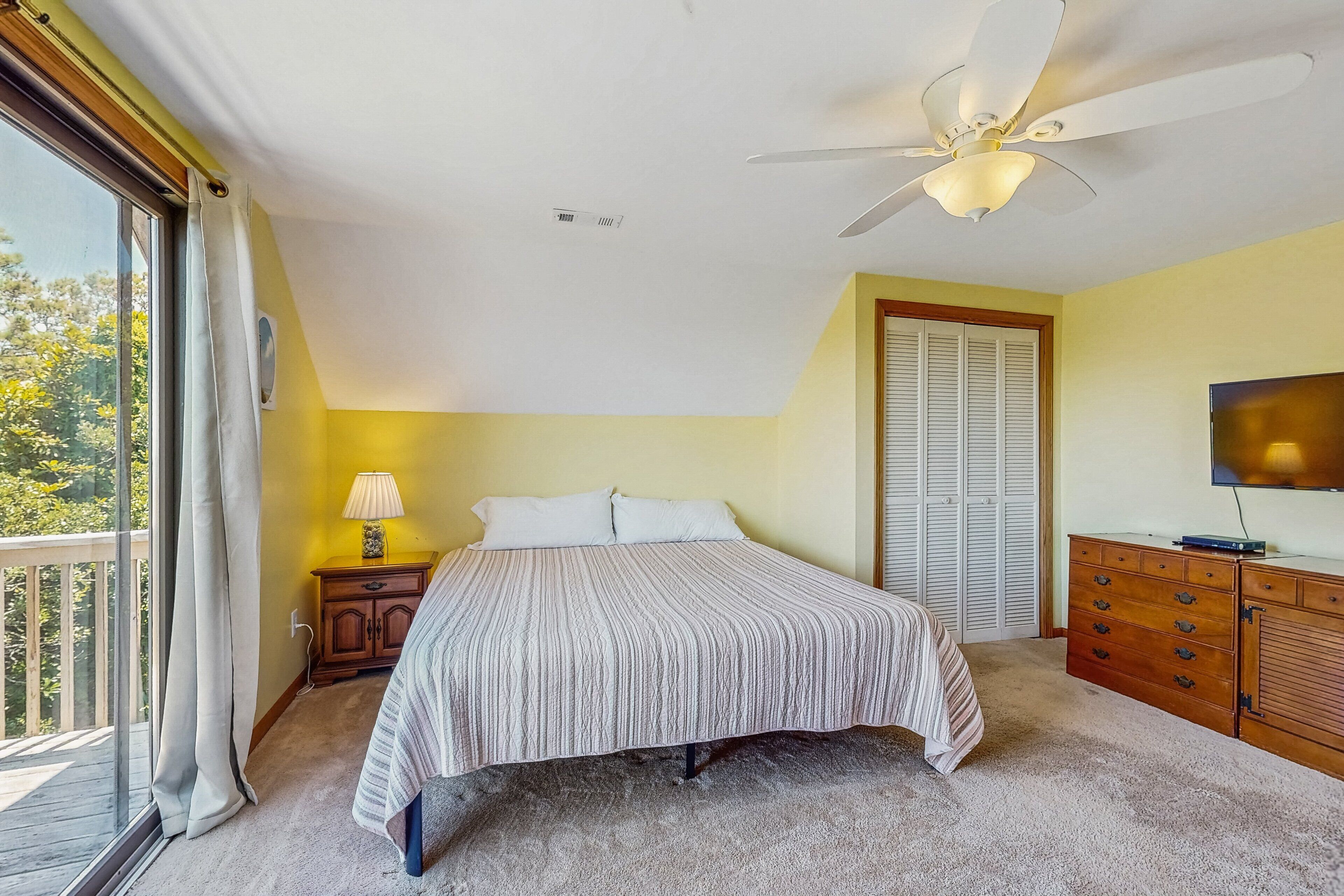 Une chambre à coucher comprenant un lit, un ventilateur de plafond, une télévision, une commode en bois et une fenêtre avec des rideaux.