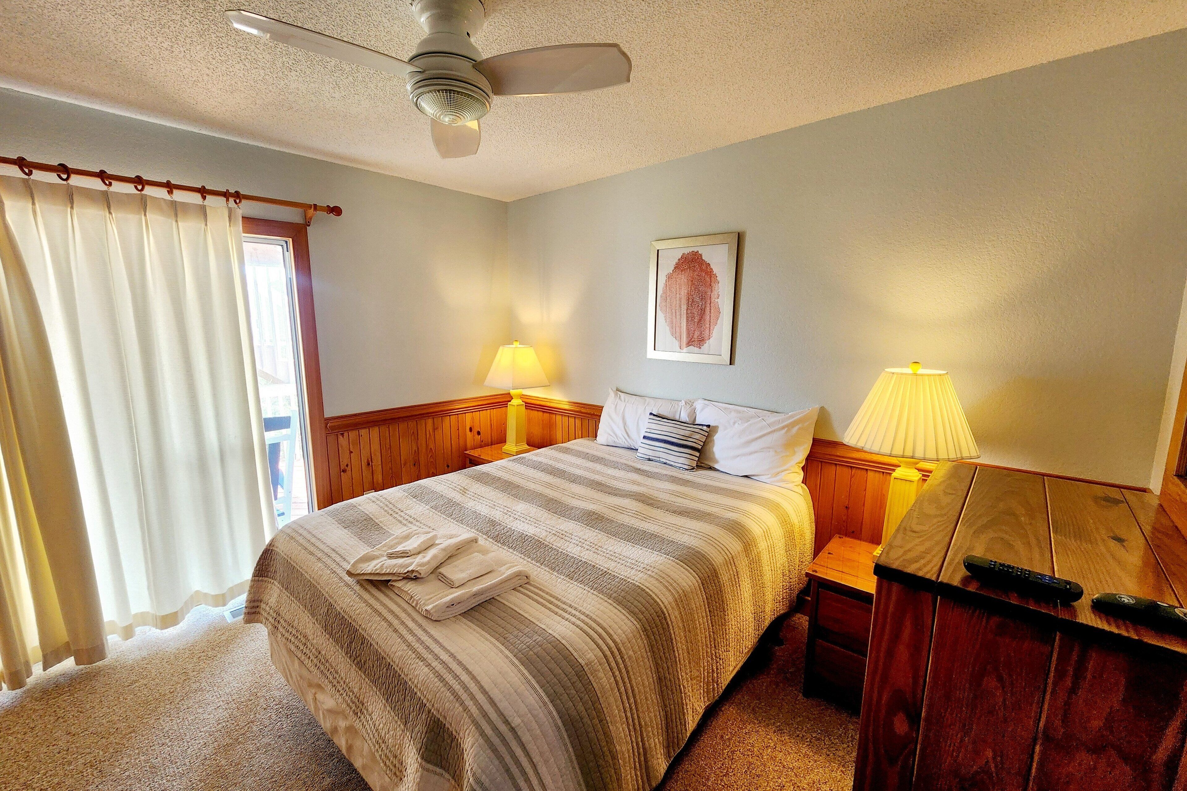 A bedroom with a bed, two bedside lamps, a ceiling fan, a window with curtains, and a framed picture on the wall.
