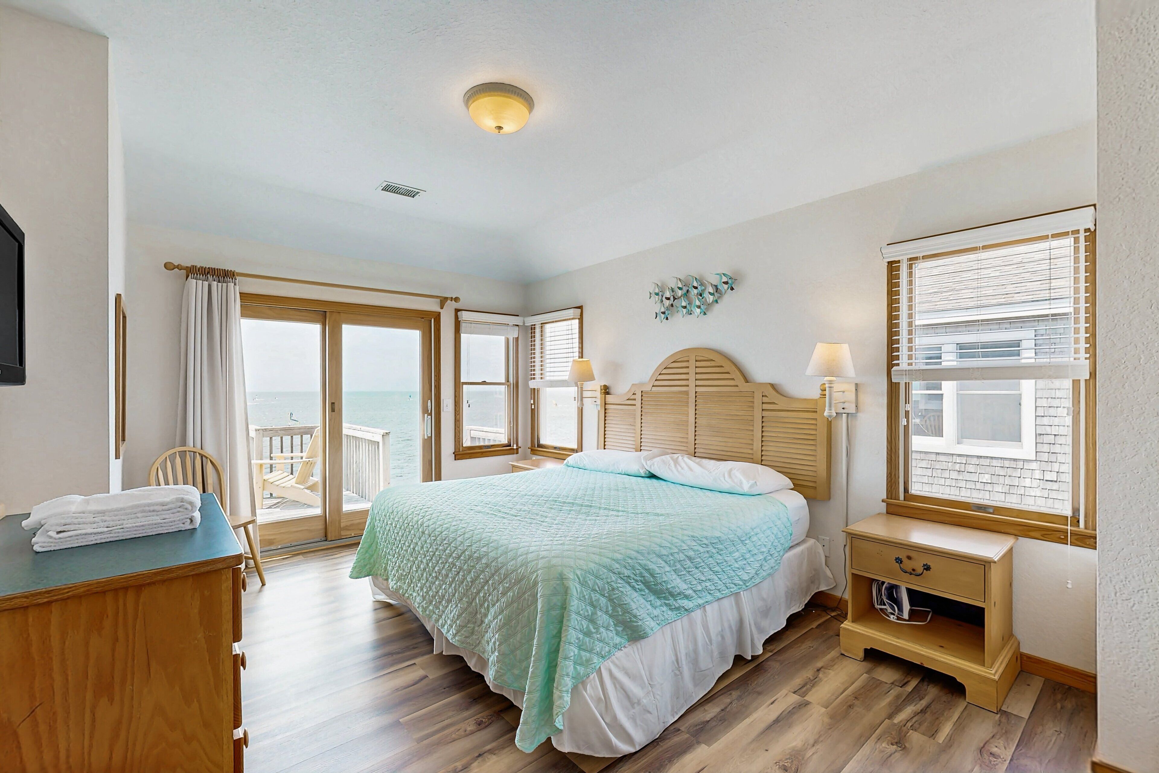 A bedroom with a bed, a wooden dresser, a nightstand, a lamp, and a view of the ocean through large windows.