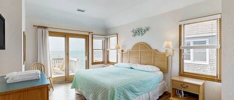 A bedroom with a bed, a wooden dresser, a nightstand, a lamp, and a view of the ocean through large windows.