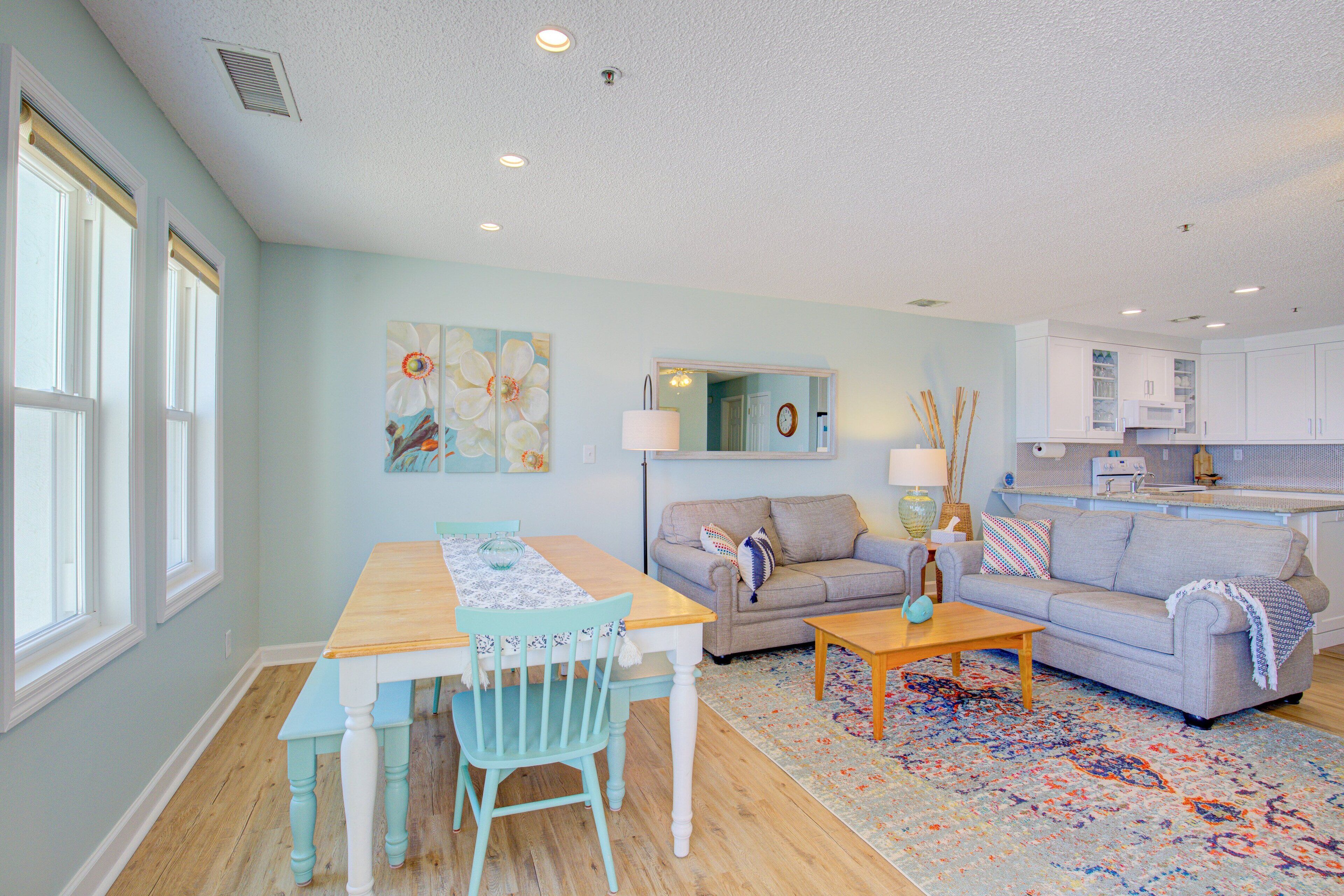 A spacious living room with a wooden dining table, a grey sofa, and a colorful rug.
