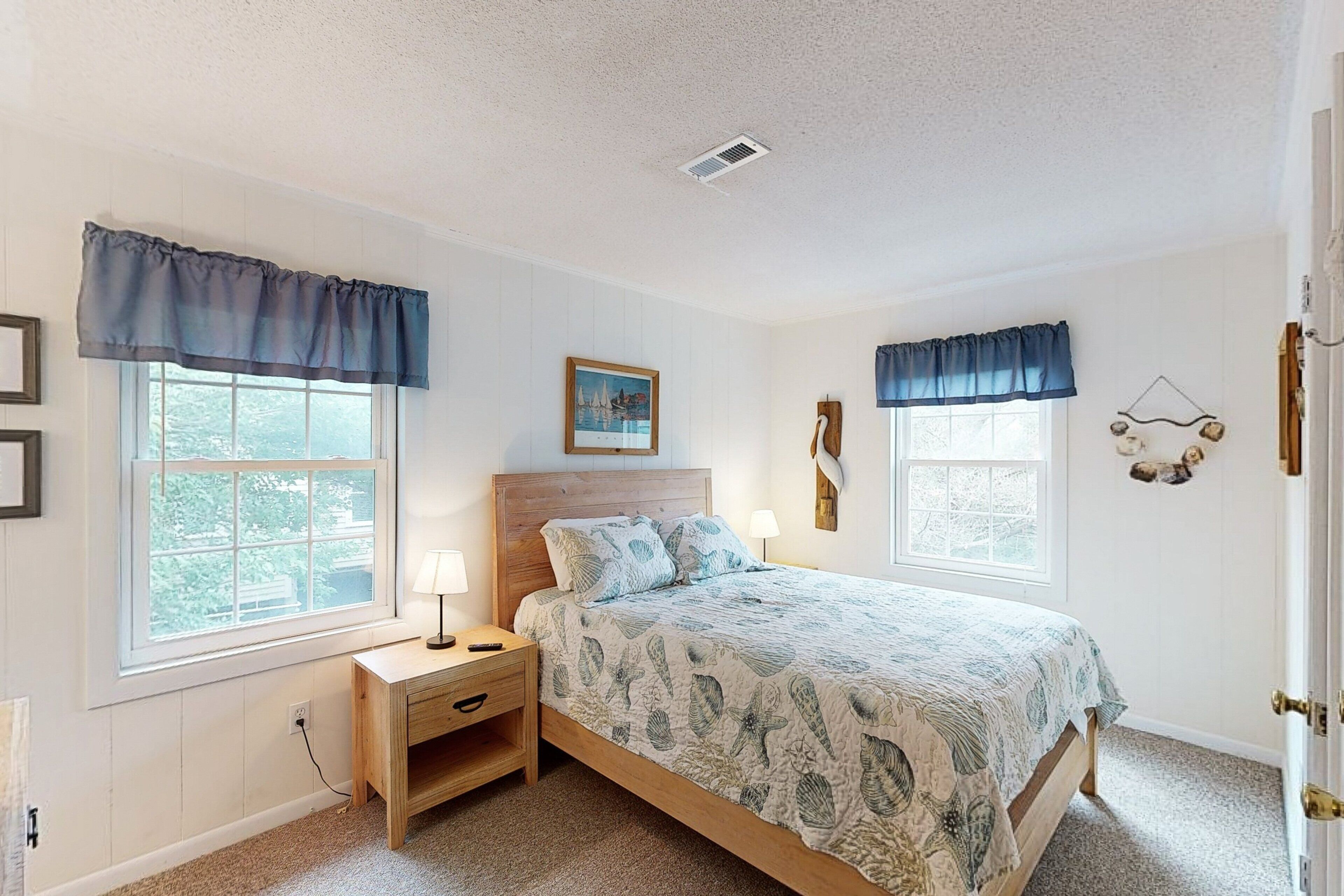 A bedroom with a wooden bed, two bedside tables, a nightstand, a window with a view, and a wall-mounted decoration.