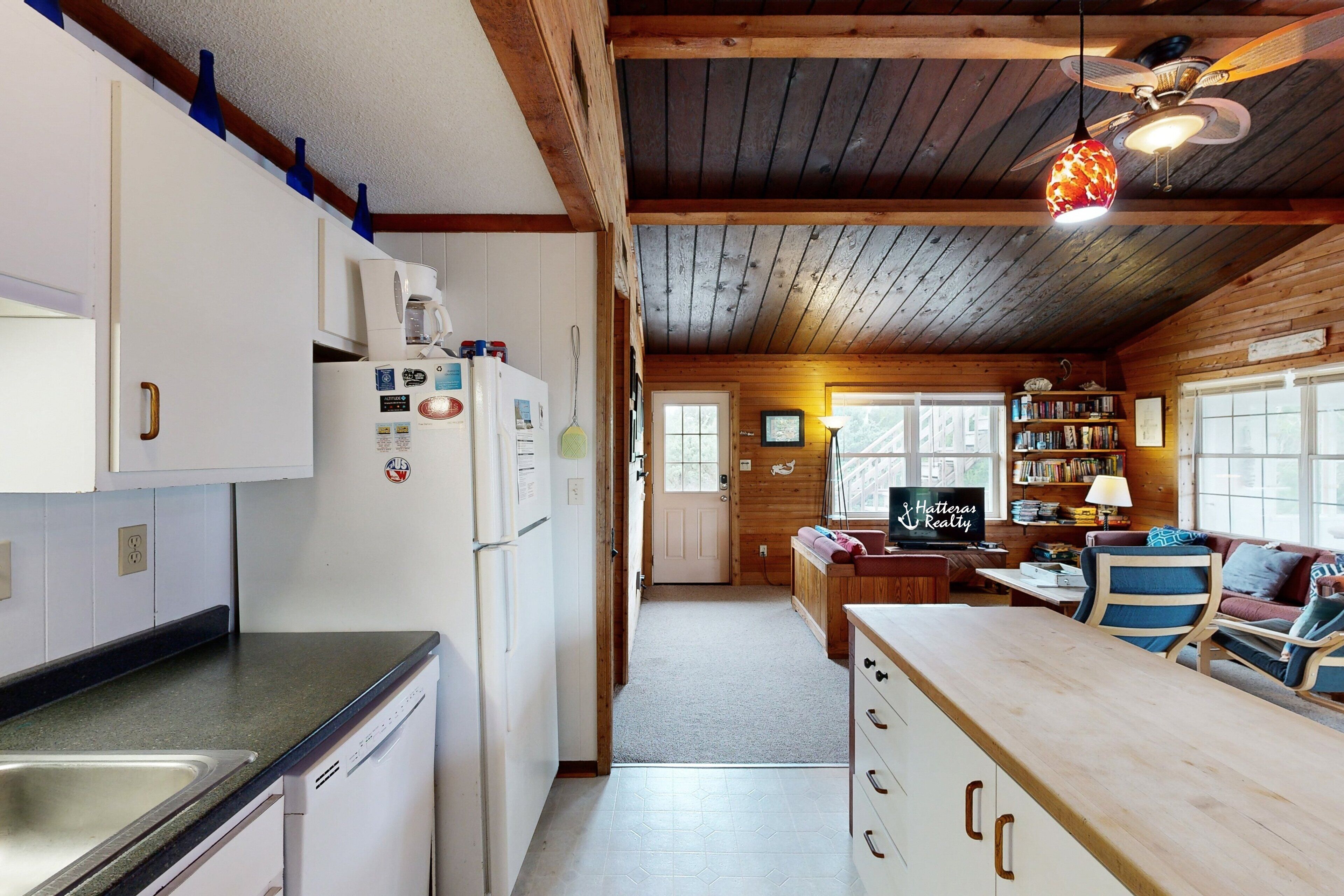 Eine kleine Küche mit Kühlschrank, Spüle und Holzschränken. Im Hintergrund ist ein Essbereich mit Tisch und Stühlen zu sehen.