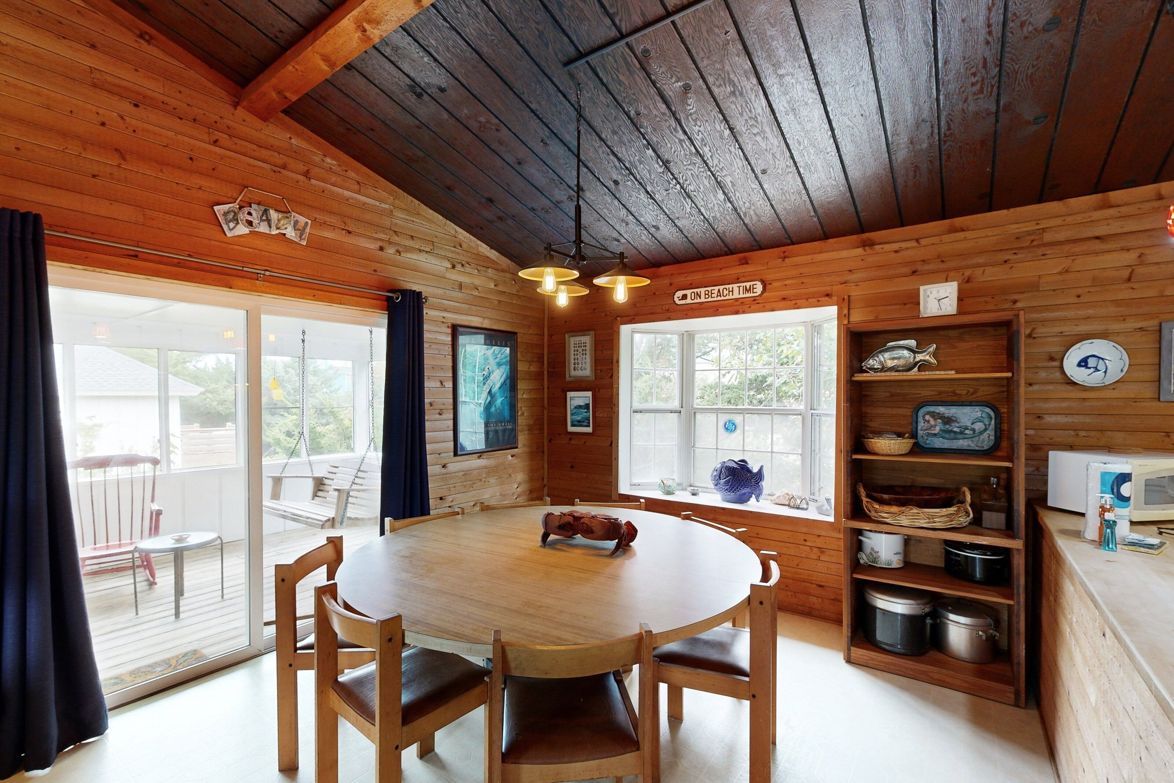 A wooden cabin interior with a round dining table, wooden chairs, a kitchen area with shelves, and a large window.