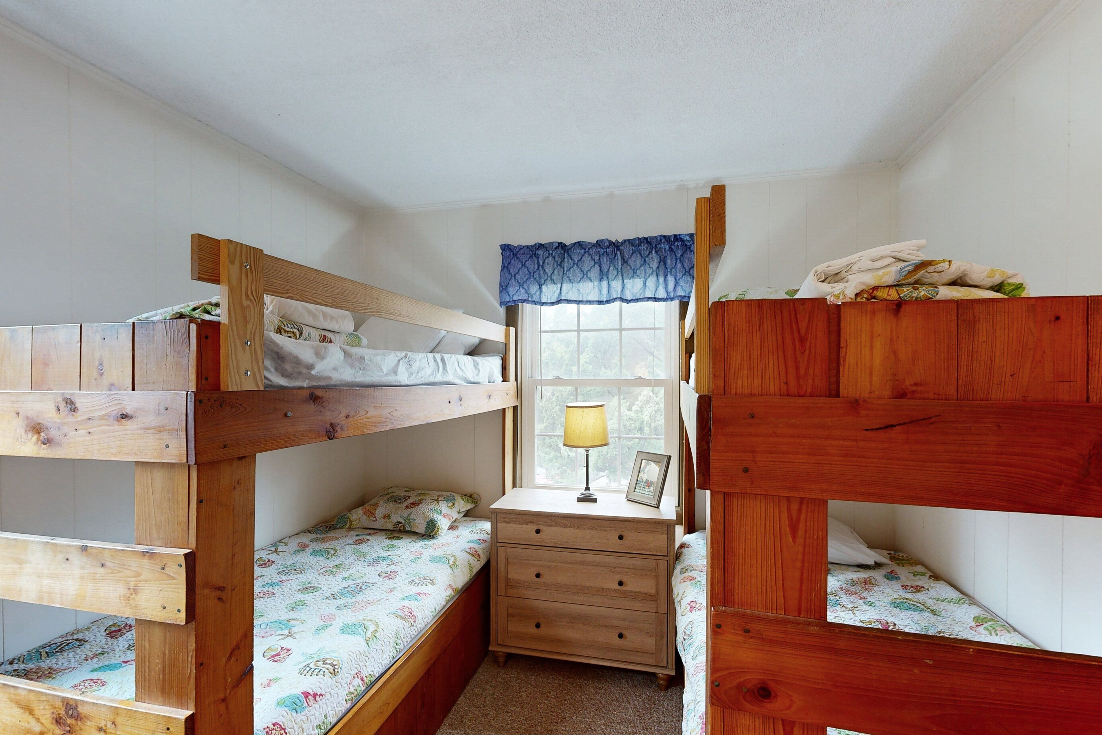 A dormitory room with two bunk beds, a small wooden desk with a lamp, and a window with a blue valance.