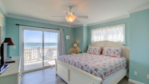 A bedroom with a bed, a ceiling fan, a nightstand, a lamp, a view of the ocean through a sliding door, and a window with curtains. - Beachfront home with beach access, elevator, ocean views, outdoor shower, W/D (North Topsail Beach)