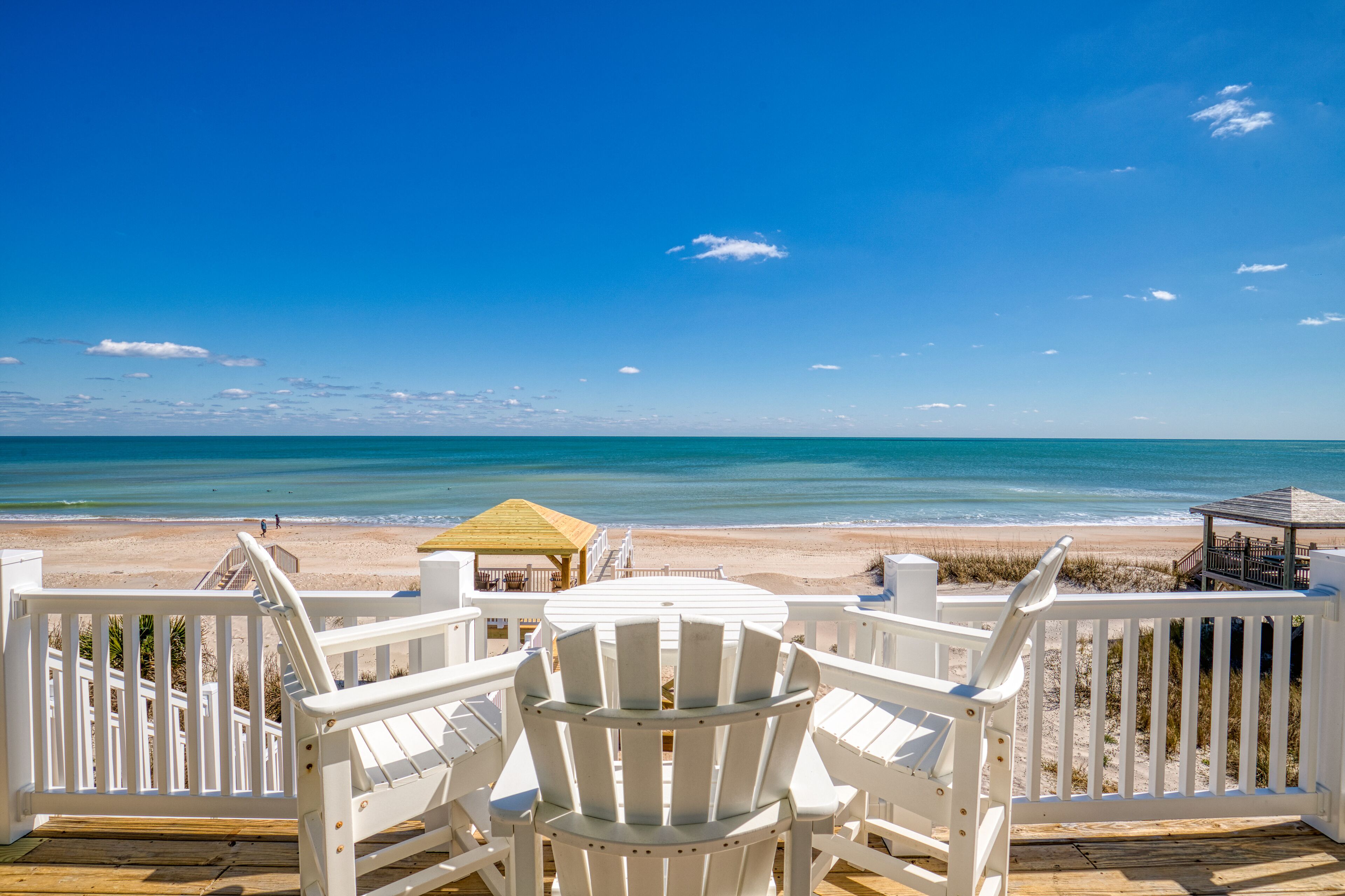A wooden deck with white Adirondack chairs overlooking a beach.