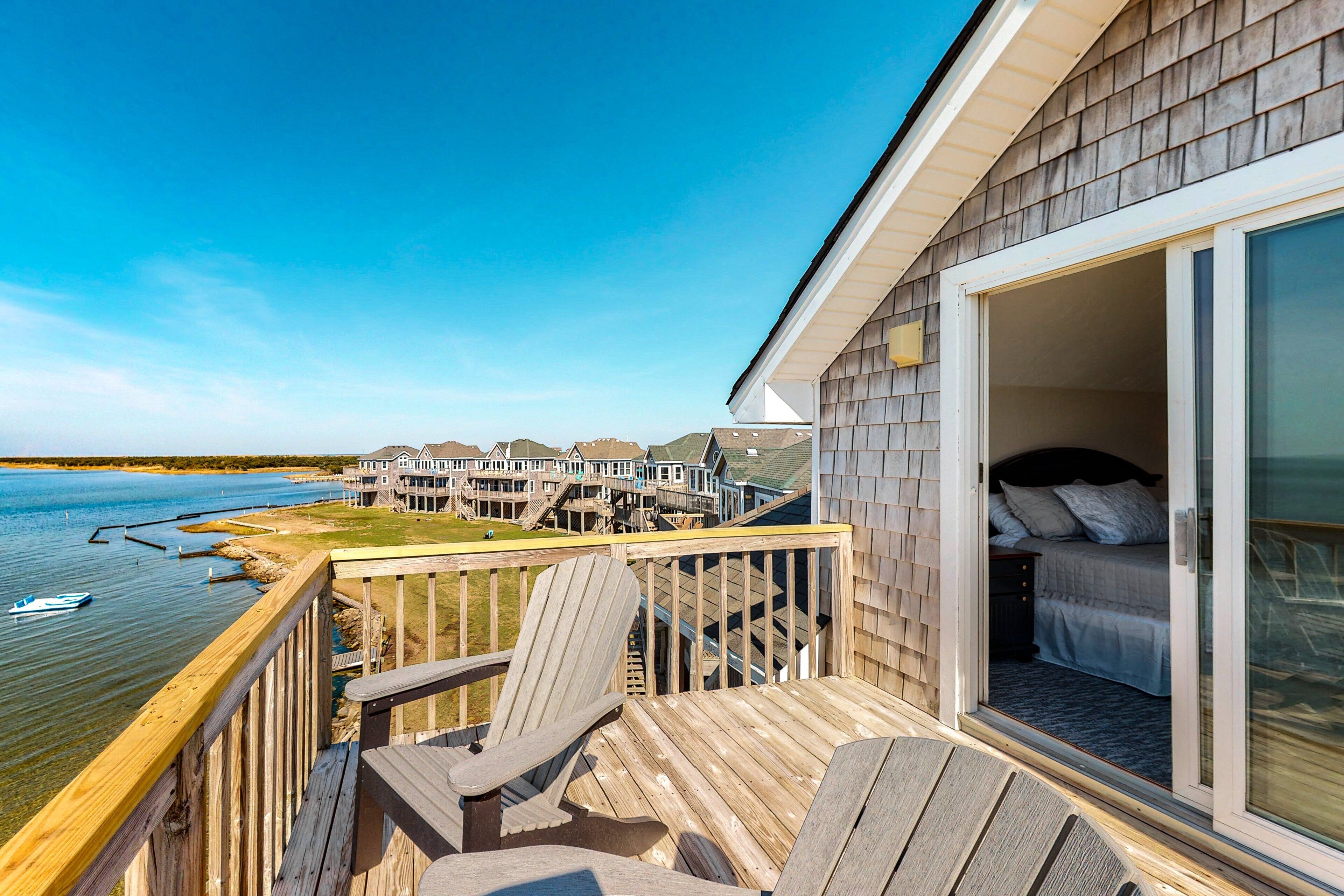 A wooden deck with chairs overlooking a body of water, a bedroom with a sliding glass door, and a view of a waterfront property.
