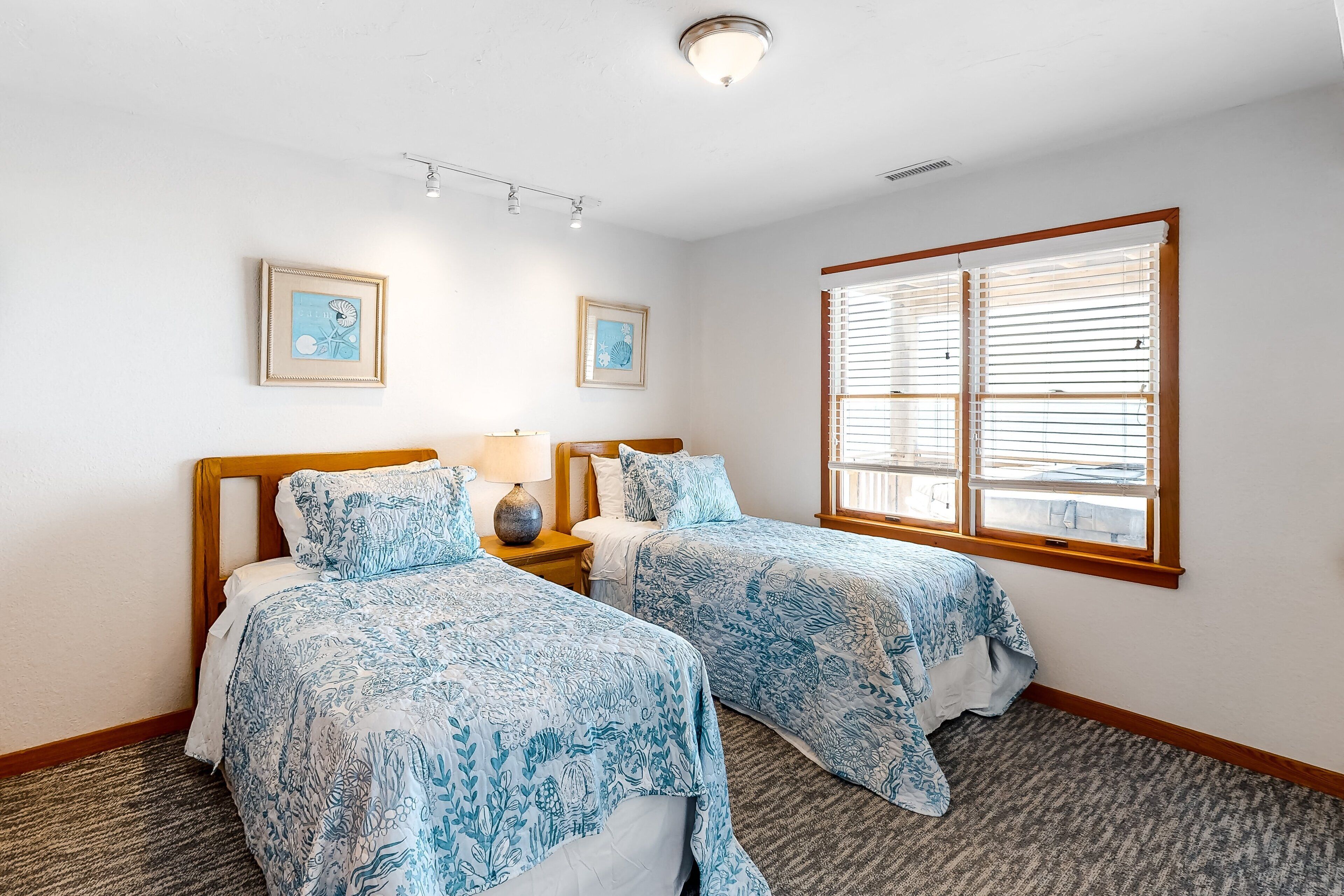 A hotel room with two beds, a window with blinds, a ceiling light, and two framed pictures on the wall.