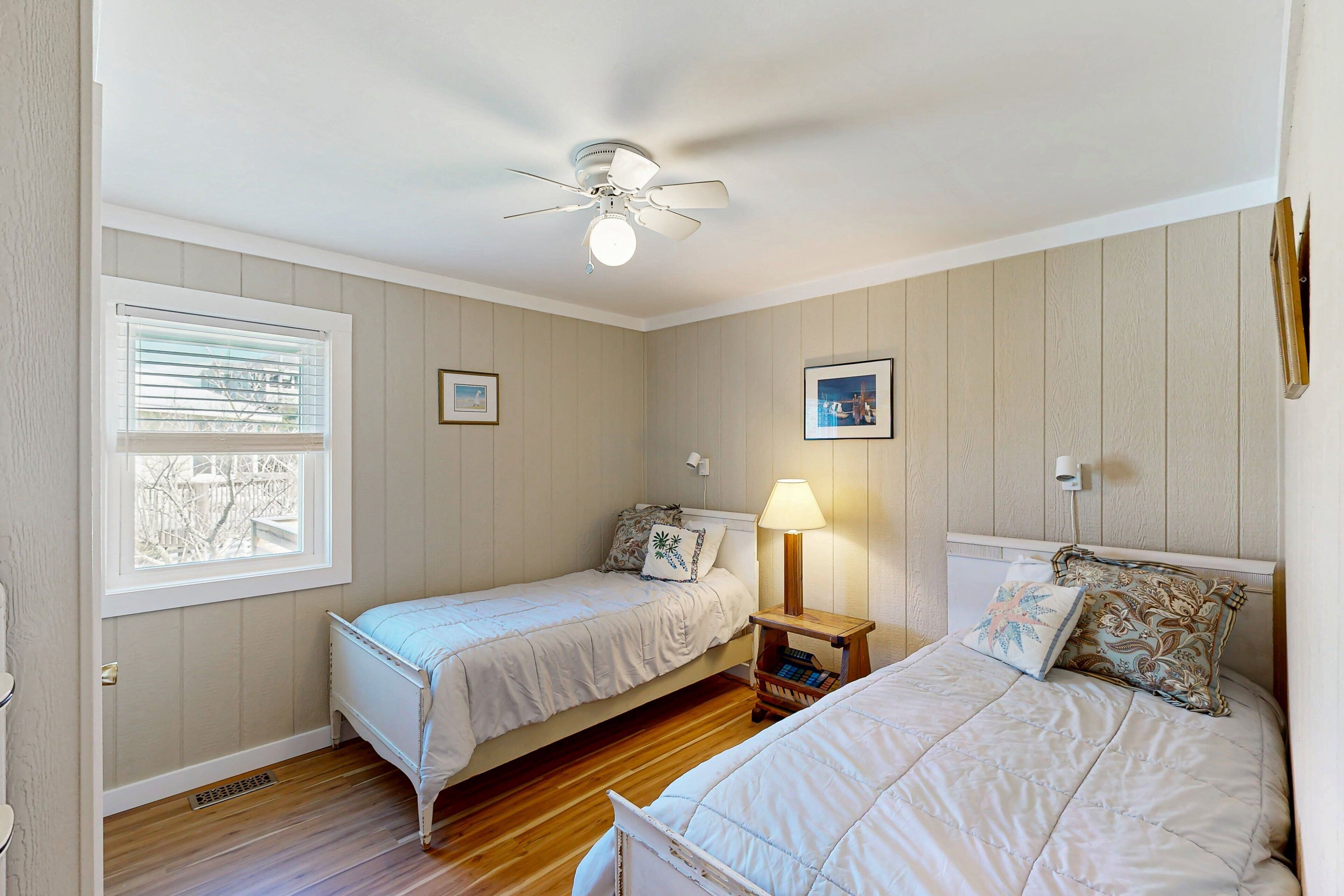 A bedroom with two single beds, a ceiling fan, a window with blinds, and a bedside table with a lamp.