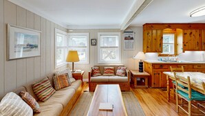 Ein gemütliches Wohnzimmer mit einer Couch, einem Couchtisch und einem Essbereich mit Tisch und Stühlen. Es gibt eine Küchenzeile mit Holzausbau und Spüle.