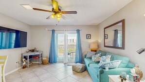 Un salon avec un canapé de couleur turquoise, une table basse blanche, un ventilateur de plafond et une porte-fenêtre coulissante donnant sur un balcon extérieur.
