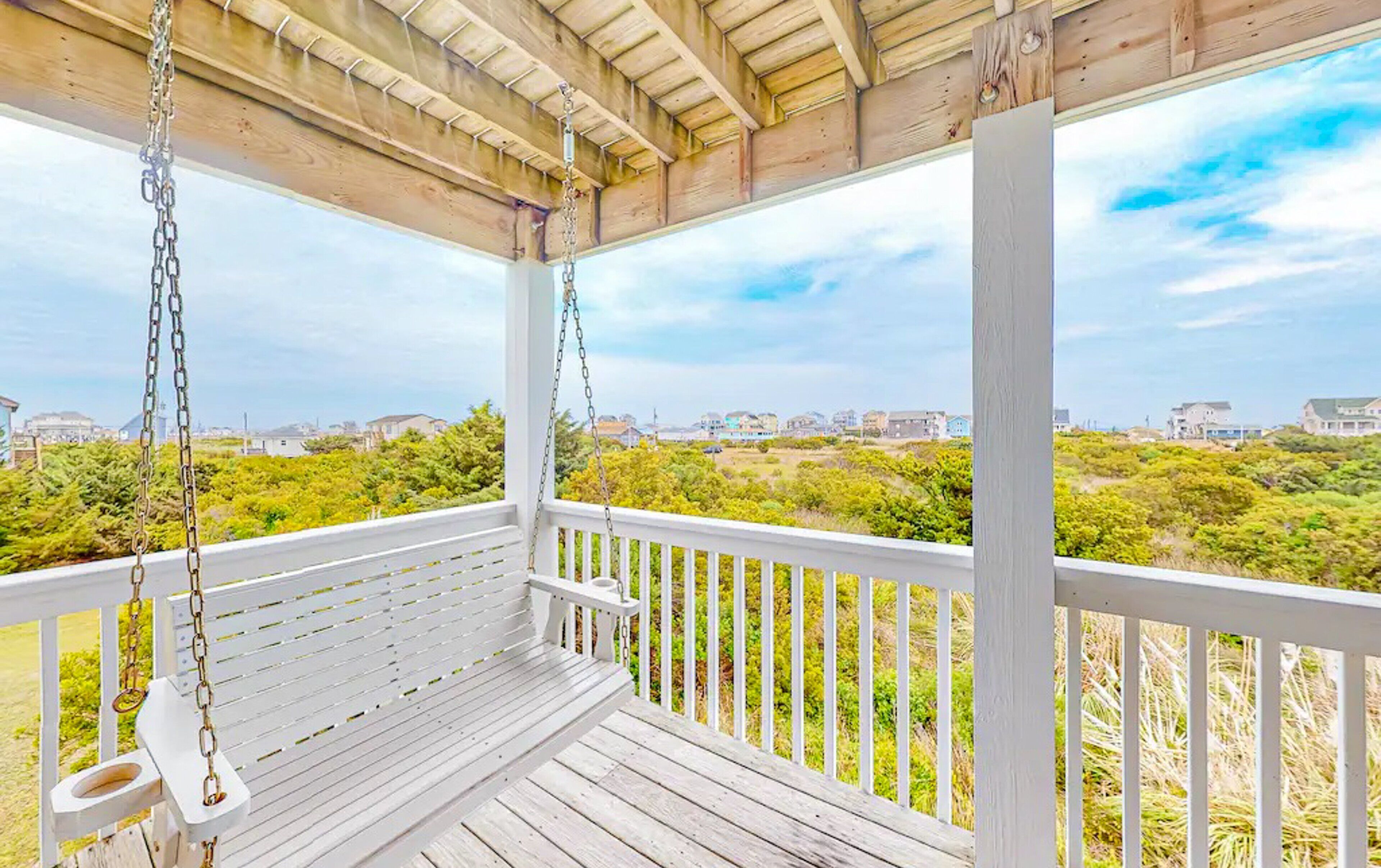 A wooden porch with a swing, white railings, and a view of a coastal landscape.