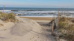 Una playa arenosa con olas acercándose a la orilla, una cerca de madera y vegetación escasa.