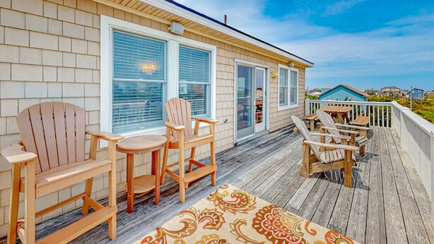 Una terraza de madera con sillas tipo Adirondack y una mesa, un porche cubierto y vistas del barrio circundante.