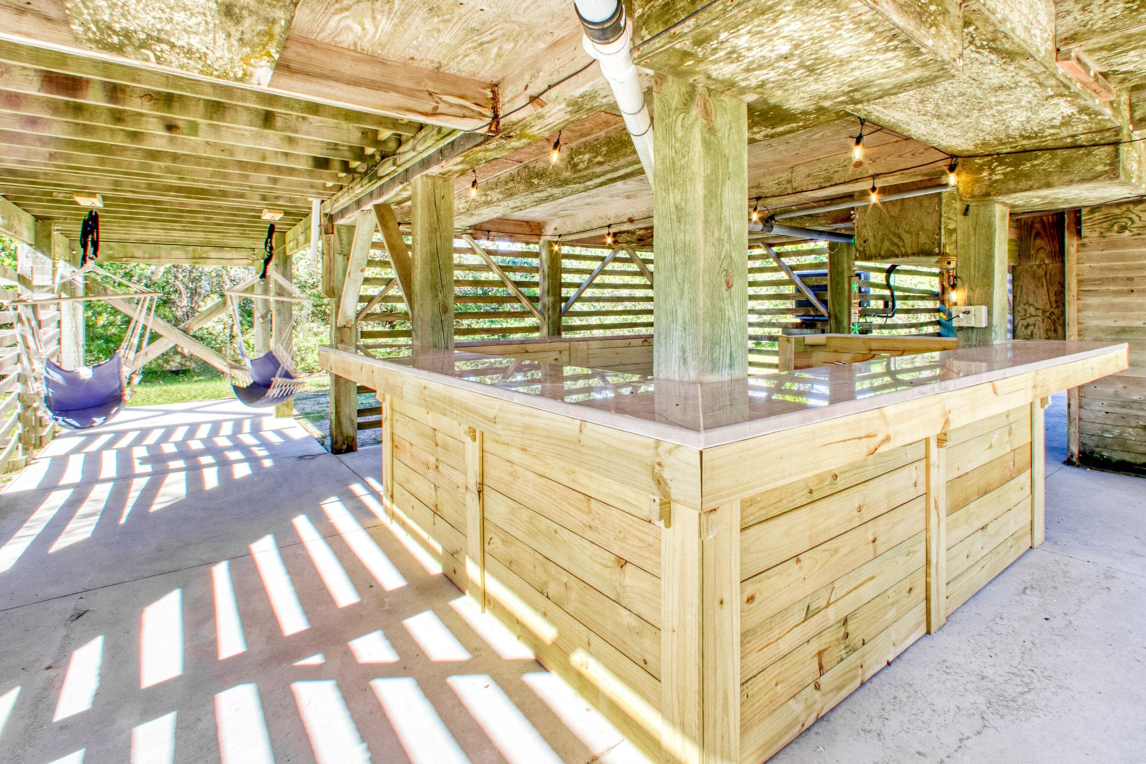 Eine Holzterrasse mit Barbereich mit Glasplatte, gestützt von Holzbalken und -säulen, mit hängenden Lichtern und Blick auf Grün im Hintergrund.