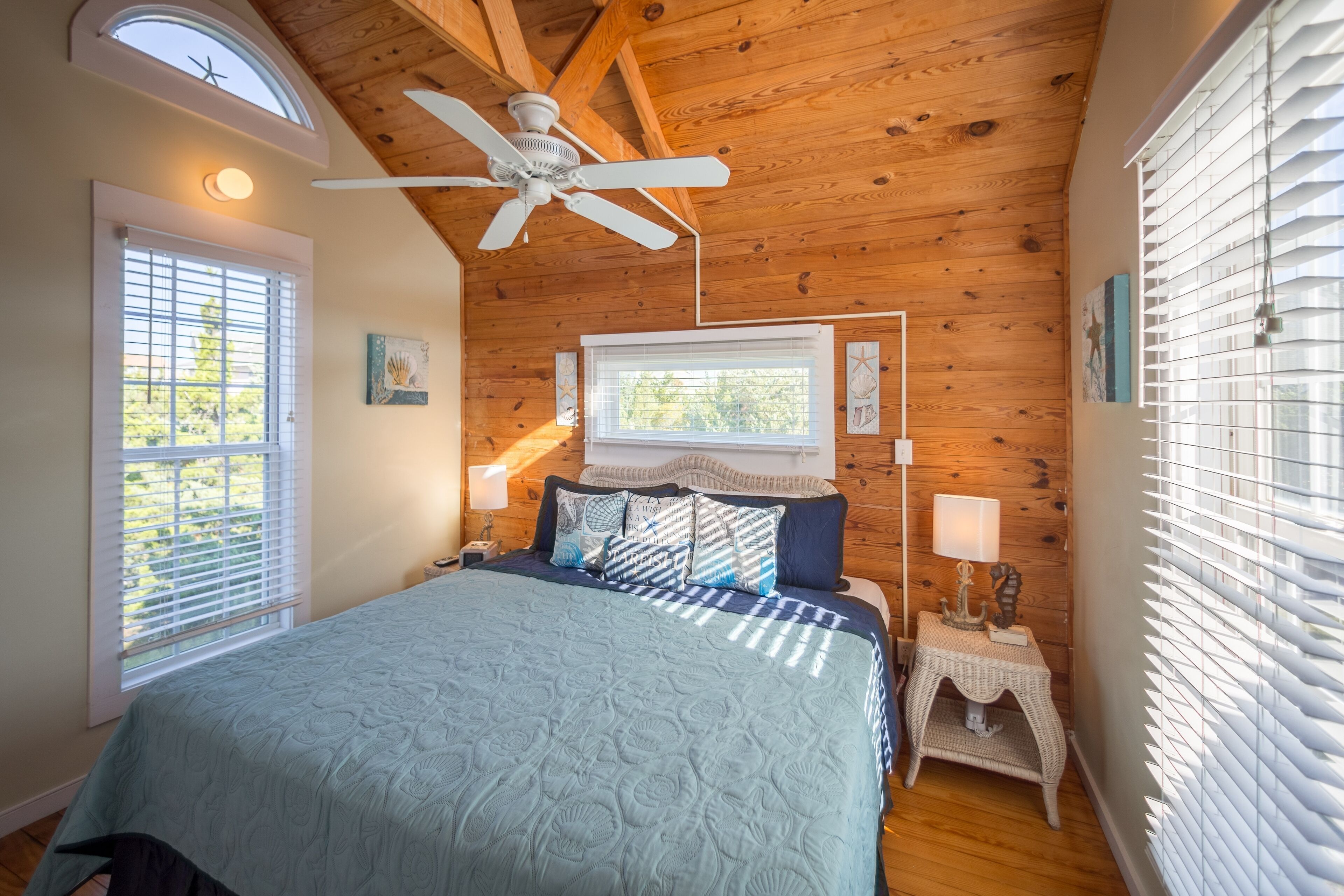 A bedroom with a wooden ceiling, a bed with a blue quilt, two bedside tables with lamps, and a window with blinds.