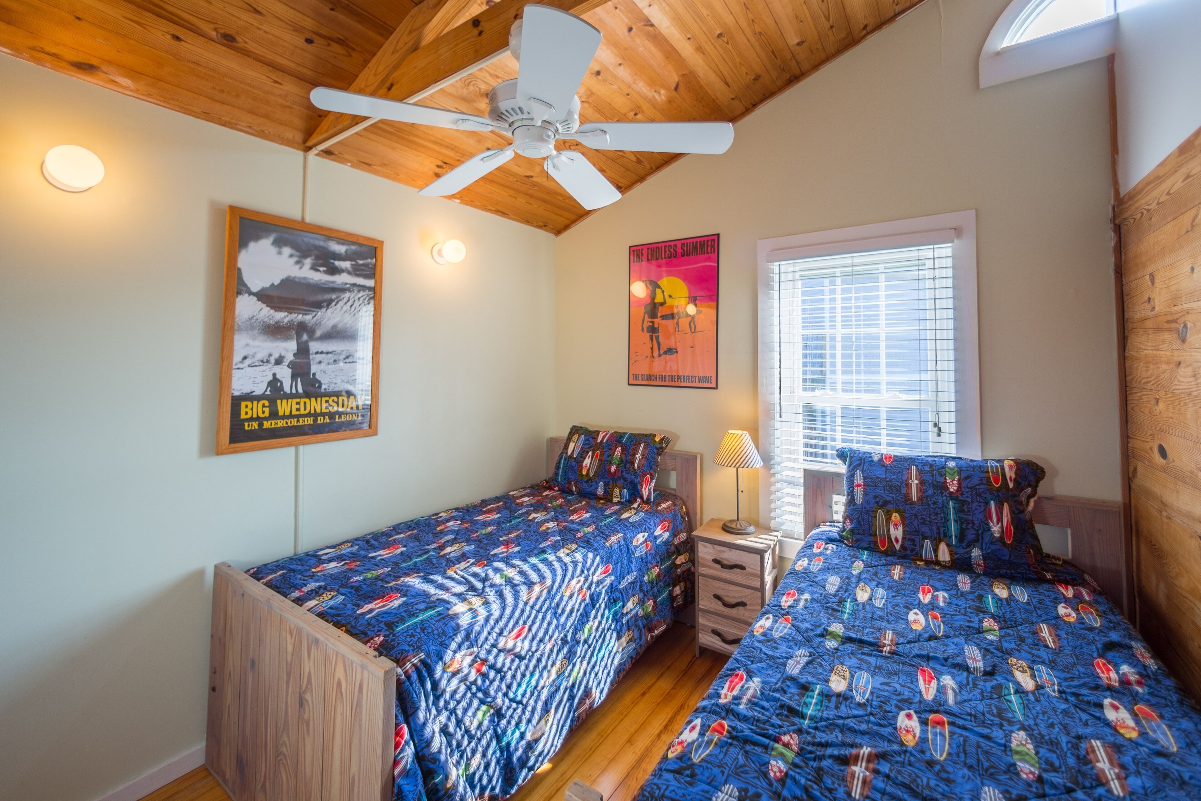 A bedroom with two beds, wooden ceiling, a ceiling fan, a poster, and a window with blinds.