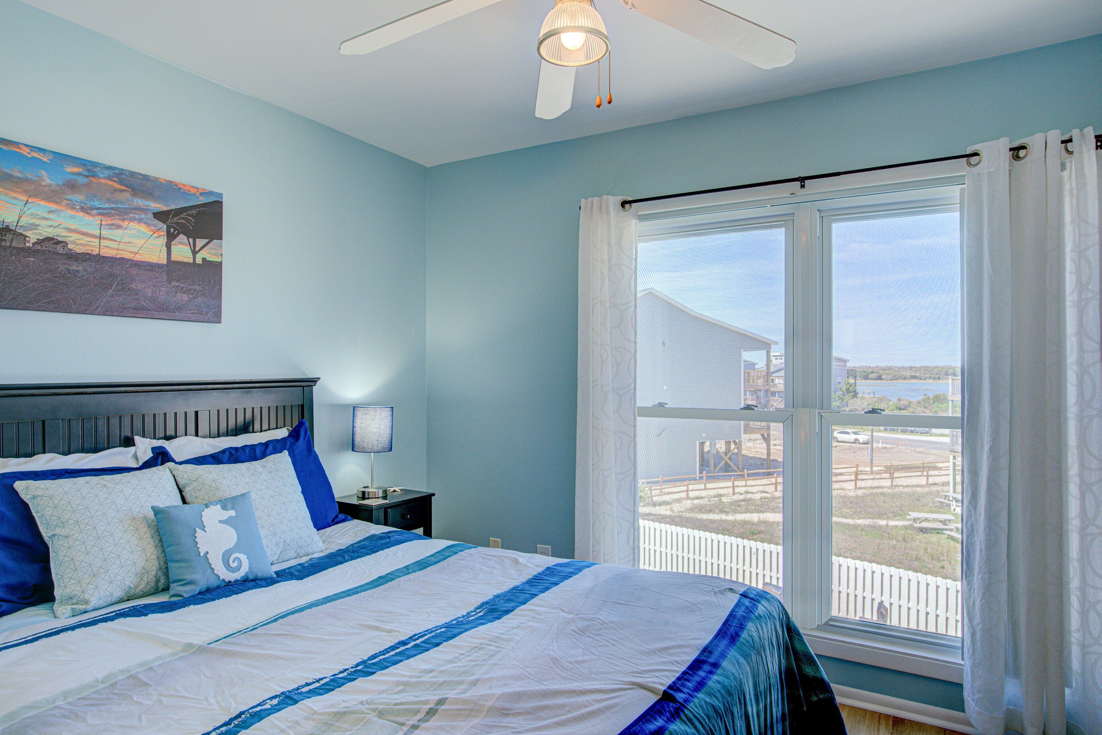 A bedroom with a bed, a nightstand, a ceiling fan, a window with curtains, and a painting on the wall.