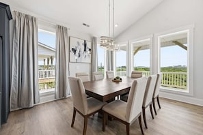 A dining area with a wooden table, six chairs, a chandelier, and large windows with a view of greenery.