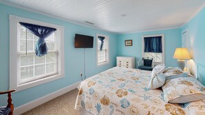 A bedroom with a bed, two windows with curtains, a dresser, a chair, a lamp, and a flat-screen TV.