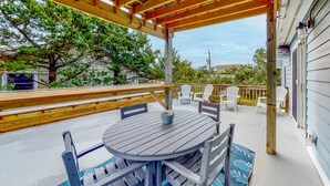 A covered outdoor patio with a round table and chairs, wooden deck, and a view of trees and buildings in the background.