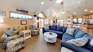 A spacious living room with a large blue sofa, a patterned armchair, and a white coffee table. There is a kitchen area with wooden cabinets and a dining table in the background. - 3-story luxury home with pool, hot tub, AC, tennis, & deck - dog-friendly (Avon)