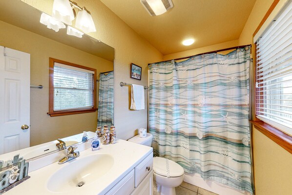 A bathroom with a white sink, mirror, and toilet. There is a window with blinds, a shower curtain with a wave pattern, and a towel rack with a white towel.