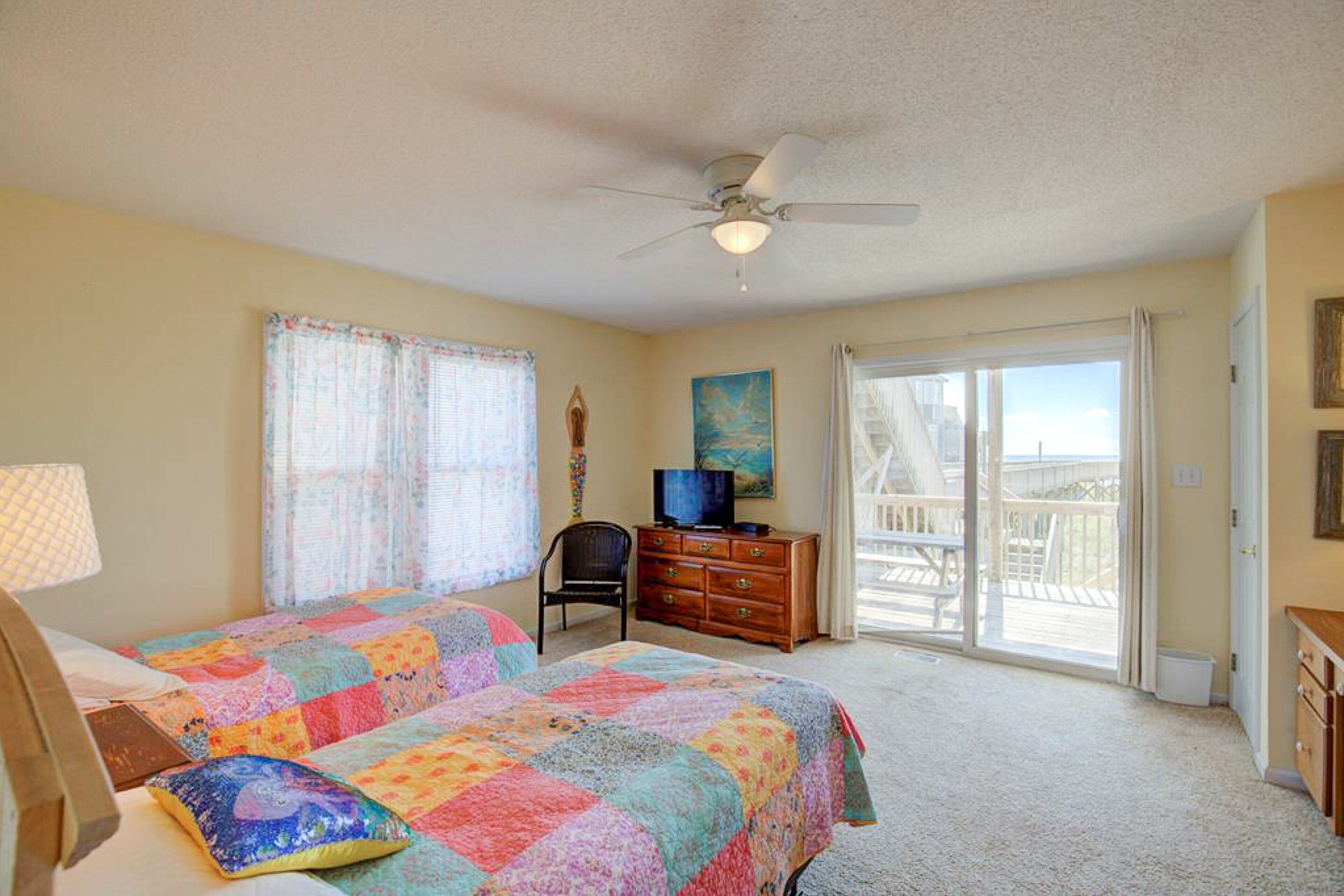 A bedroom with two beds, a ceiling fan, a television, and a view of the outdoors through a sliding glass door.