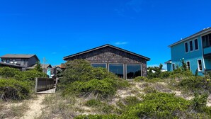 Una casa frente a la playa con un amplio ventanal corredero, rodeada de vegetación exuberante y otras viviendas.