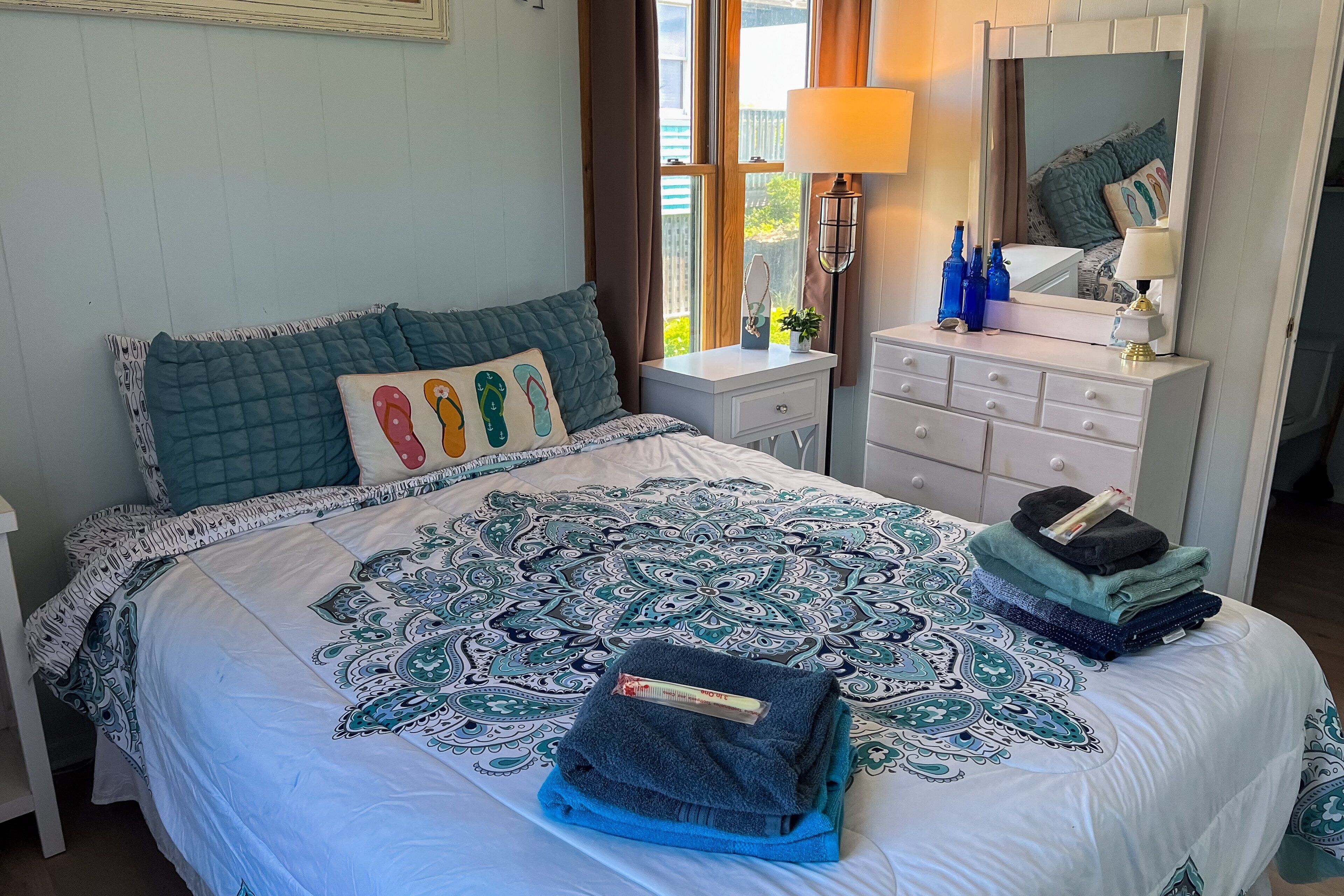 A neatly made bed with a patterned bedspread, a bedside table with a lamp, a dresser with a mirror, and a window with curtains.