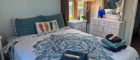 A neatly made bed with a patterned bedspread, a bedside table with a lamp, a dresser with a mirror, and a window with curtains.
