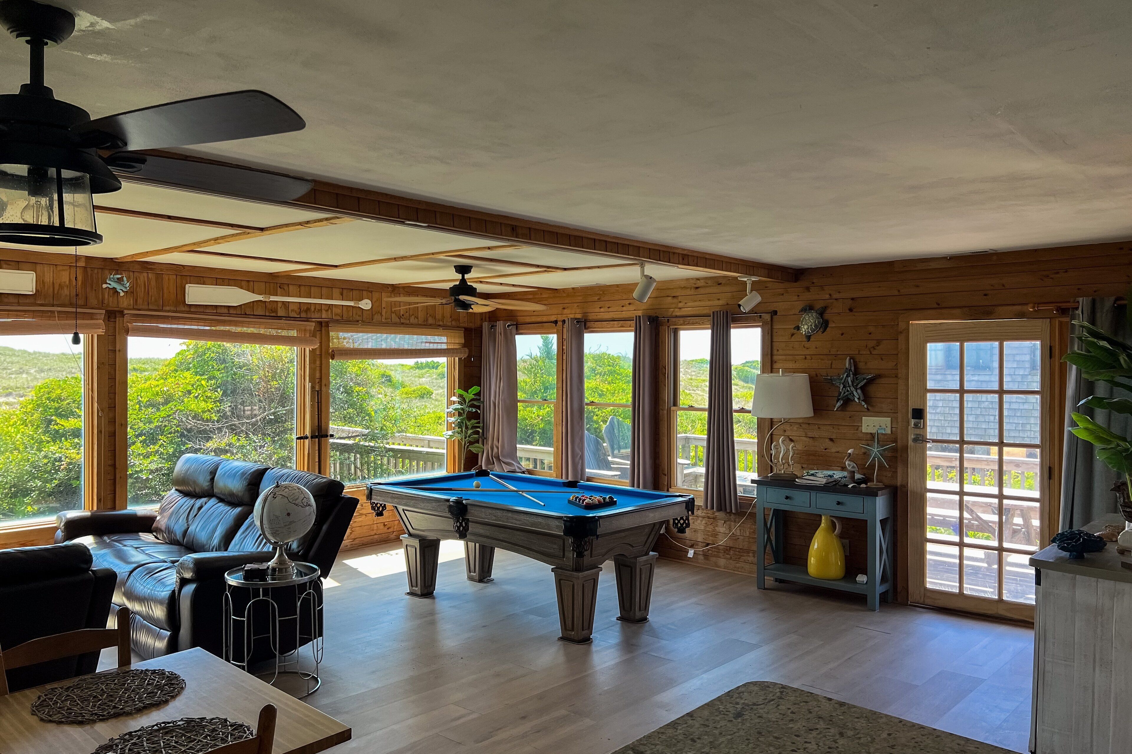 A spacious room with a pool table, leather sofa, and large windows offering a view of greenery.