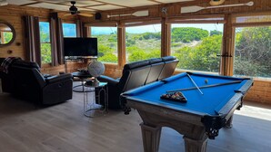 Una mesa de billar con superficie de fieltro azul, un sofá de cuero negro y un televisor sobre un soporte.