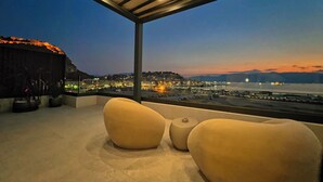 Eine Dachterrasse mit Blick auf eine Stadtlandschaft im Abenddämmerung, ausgestattet mit einer modernen Lounge mit zwei großen, runden, beige farbenen Sesseln und einem kleinen Tisch.