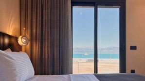 Ein Hotelzimmer mit einem Bett, einem Vorhang, einer Wandleuchte und einem großen Fenster mit Blick auf Strand und Berge.