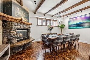 Un coin repas agrémenté d’une cheminée en pierre, de poutres en bois et d’une table dressée pour six personnes, avec des chaises à carreaux.