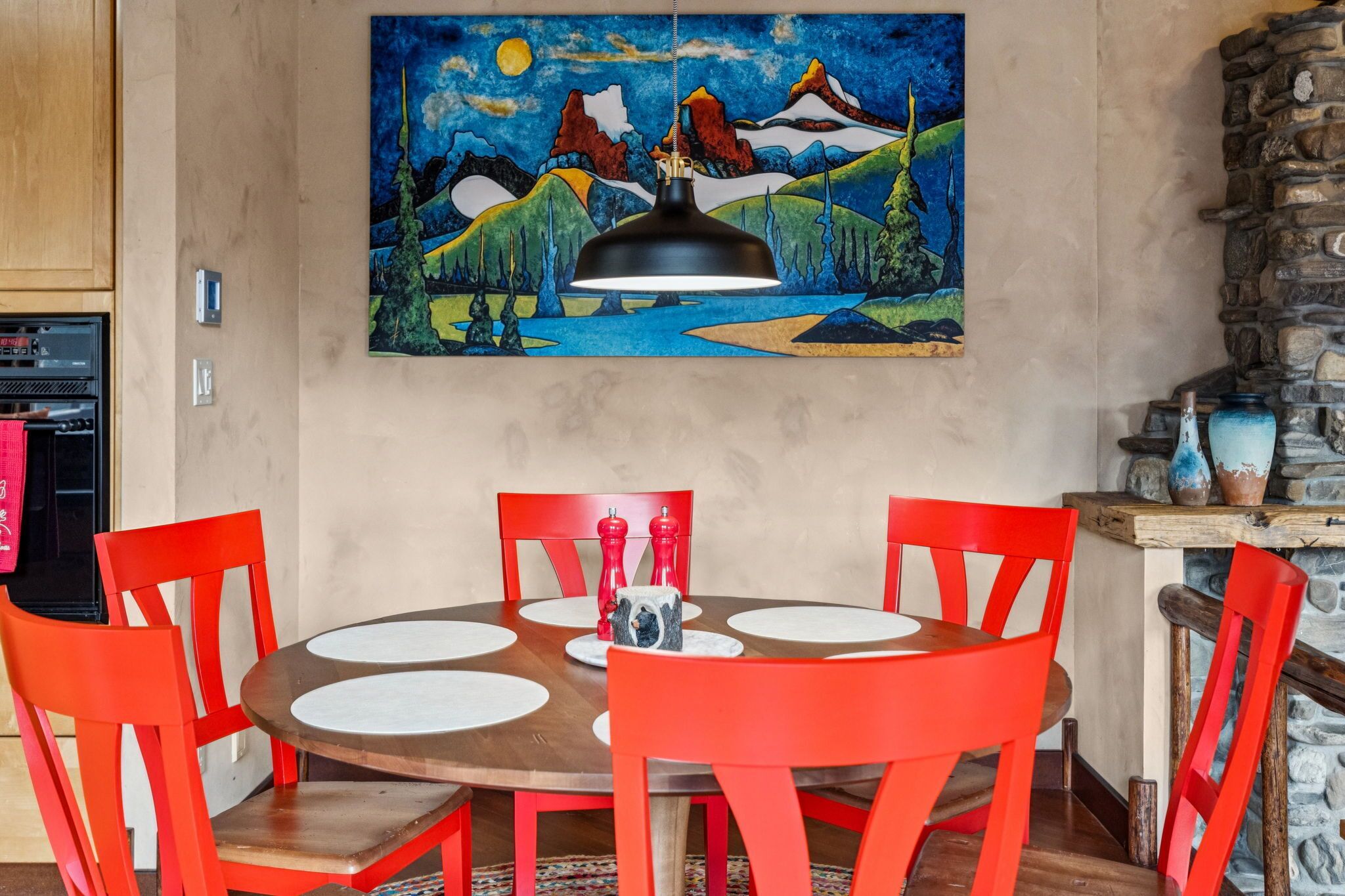 A dining area with a round table, red chairs, a painting on the wall, and a stone wall.