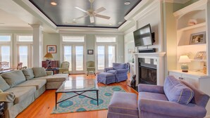 A spacious living room with a ceiling fan, a fireplace, and a television mounted above it. - Beachfront home with private heated pool, hot tub, & elevator (North Topsail Beach)