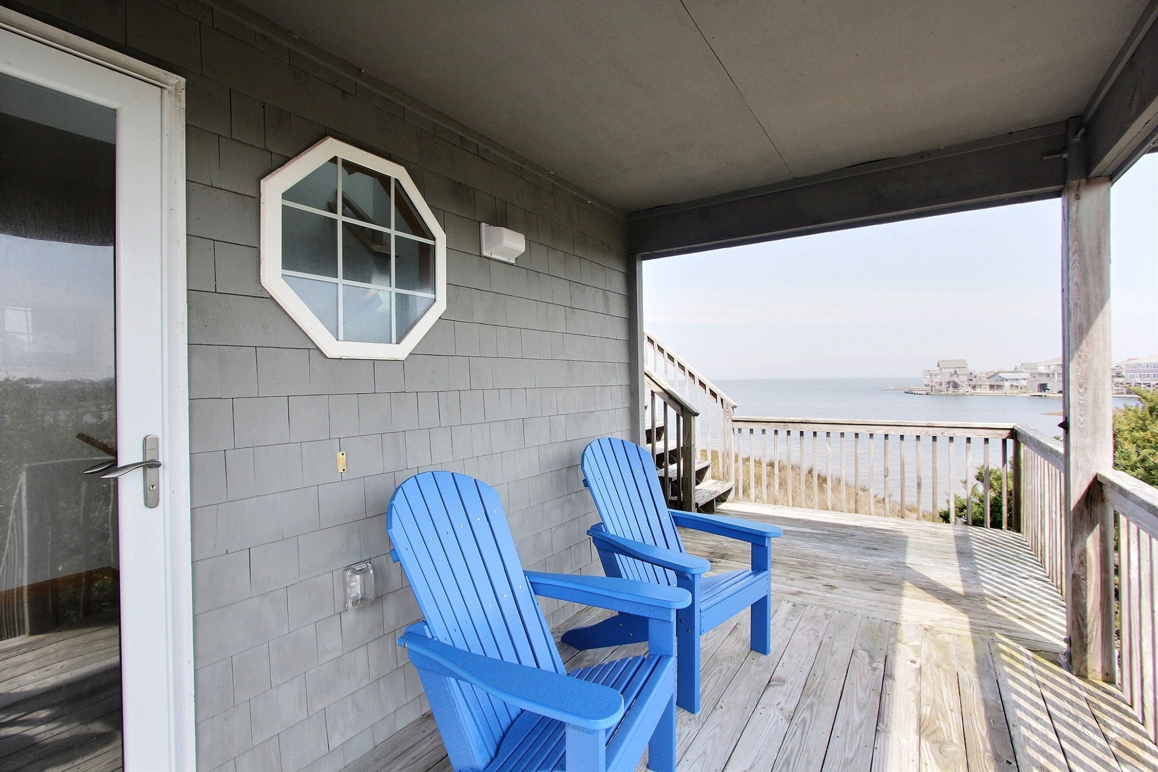 Un porche cubierto con dos sillas Adirondack azules, un ventanal hexagonal y vistas al océano.