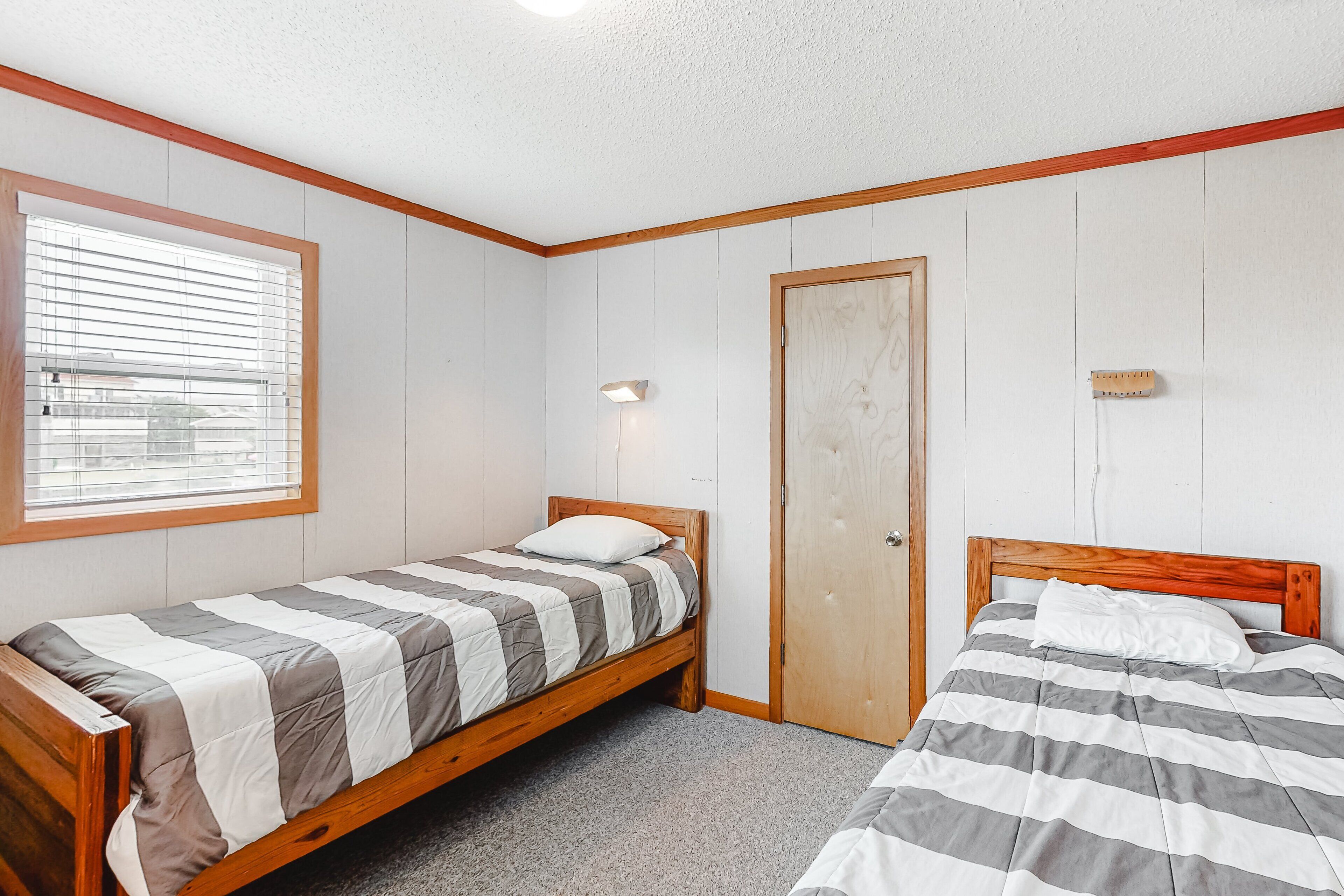 Une chambre avec deux lits simples, une fenêtre avec des stores, une porte et un luminaire fixé au mur.