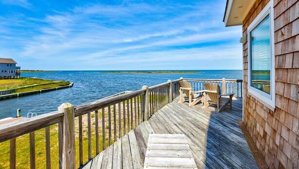 A wooden deck with a railing overlooking a body of water, a small building, and a distant shoreline.