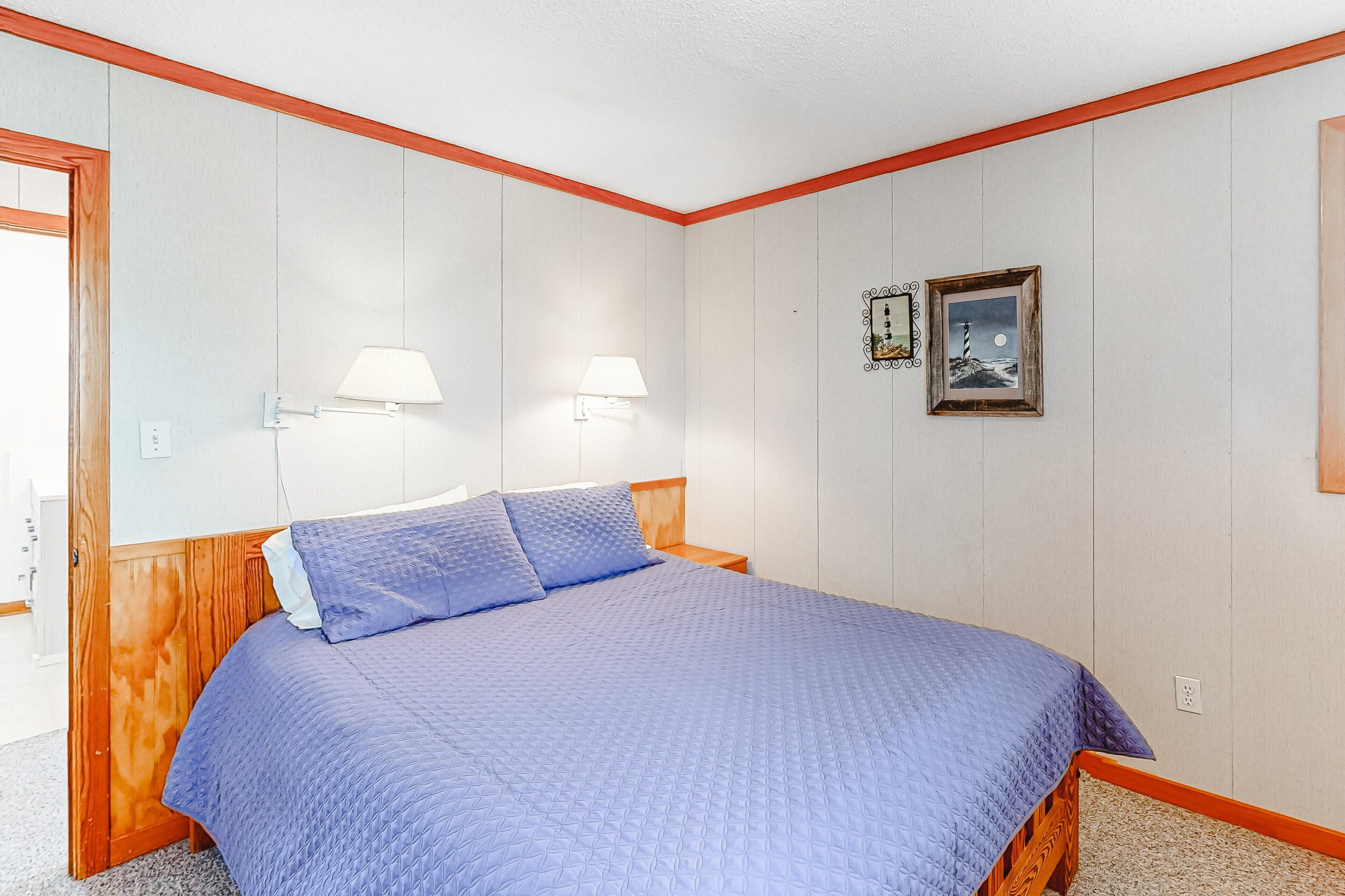 A bedroom with a wooden headboard, a blue quilted bedspread, two wall-mounted lamps, and a framed picture on the wall.