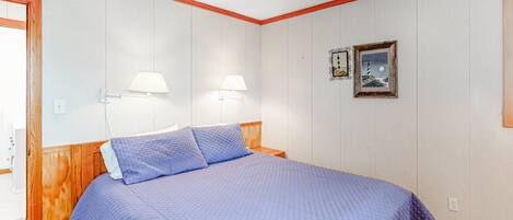 A bedroom with a wooden headboard, a blue quilted bedspread, two wall-mounted lamps, and a framed picture on the wall.